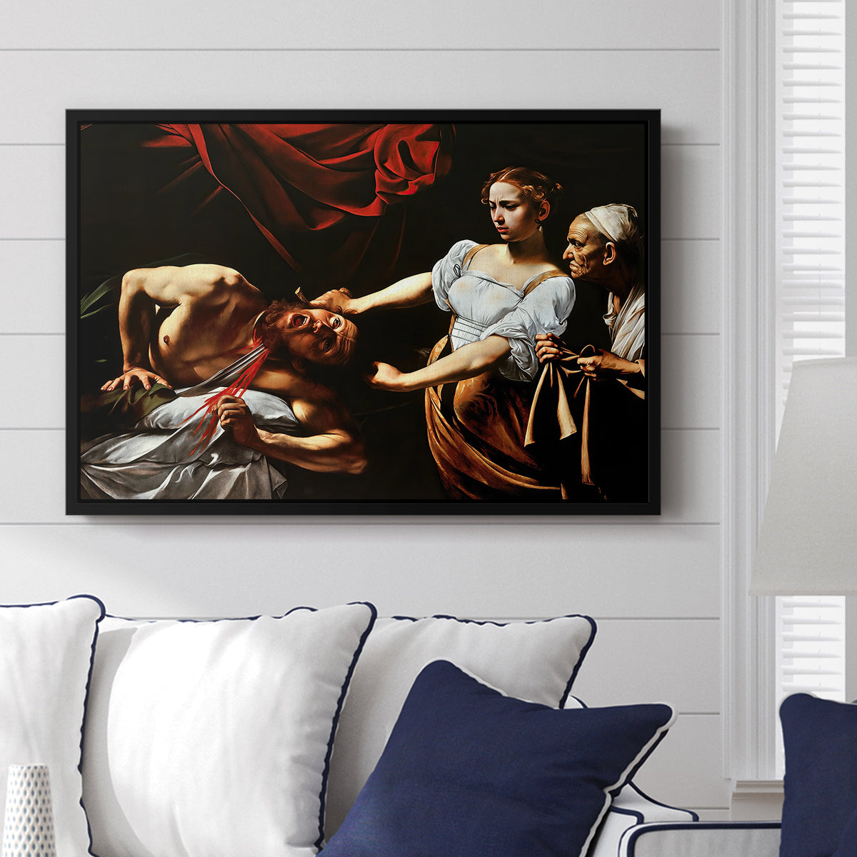 Judith Beheading Holofernes, c. 1599 by Caravaggio displayed above a sofa in a living room in a black floater frame, featuring a dramatic biblical scene of woman beheading man in bed.