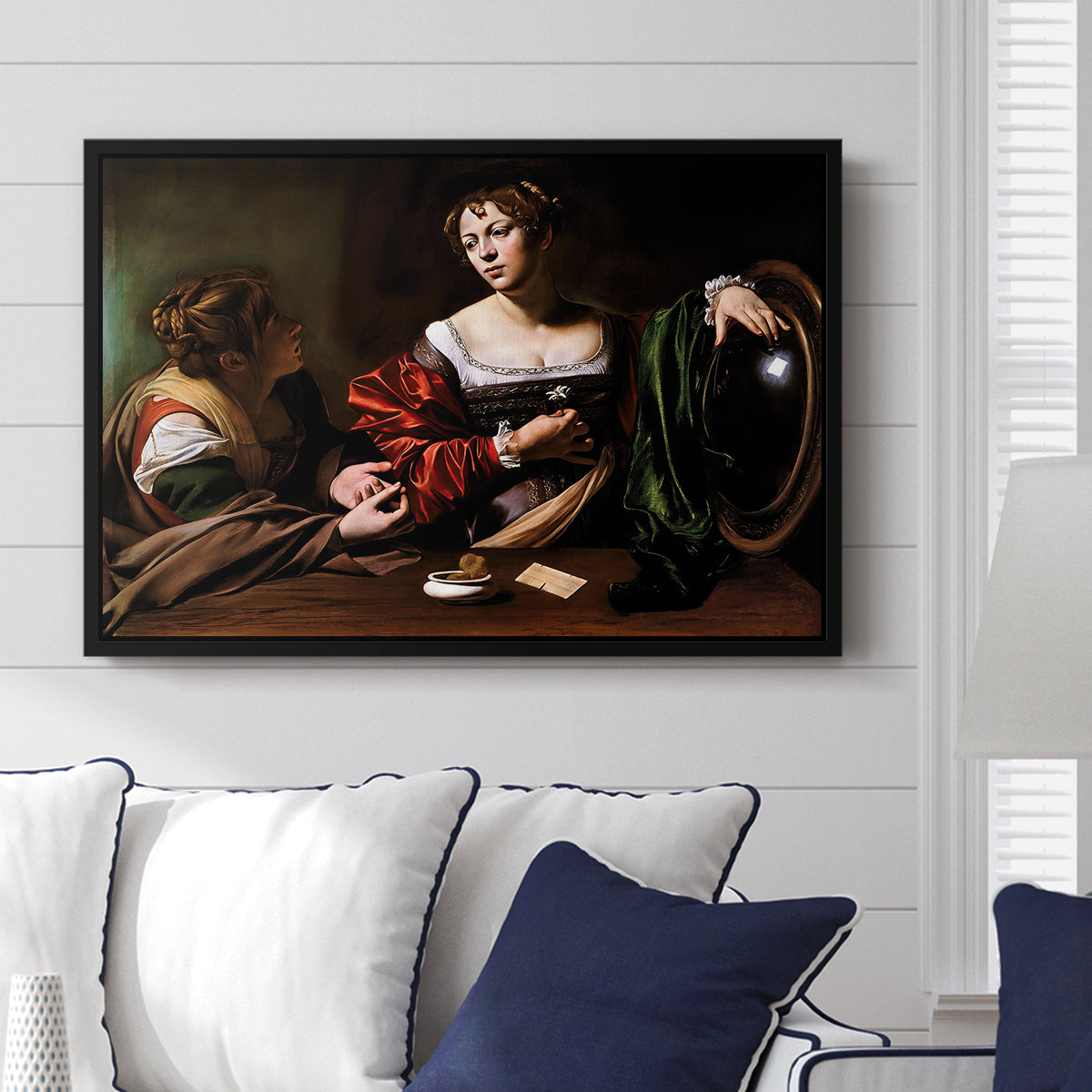 Martha and Mary Magdalene, c. 1598 by Caravaggio displayed above a sofa in a living room in a black floater frame, featuring a biblical scene of Martha speaking to Mary Magdalene indoors.