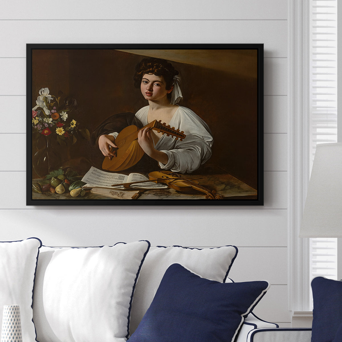 The Lute Player (Hermitage Version), c. 1596 by Caravaggio displayed above a sofa in a living room in a black floater frame, featuring a portrait of lute player surrounded by still life arrangement.