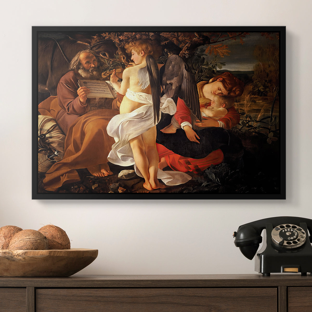 Rest on the Flight into Egypt, c. 1597 by Caravaggio in a black floater framed canvas displayed above a console table in a hallway, featuring a Baroque landscape with angel playing music for resting travelers.