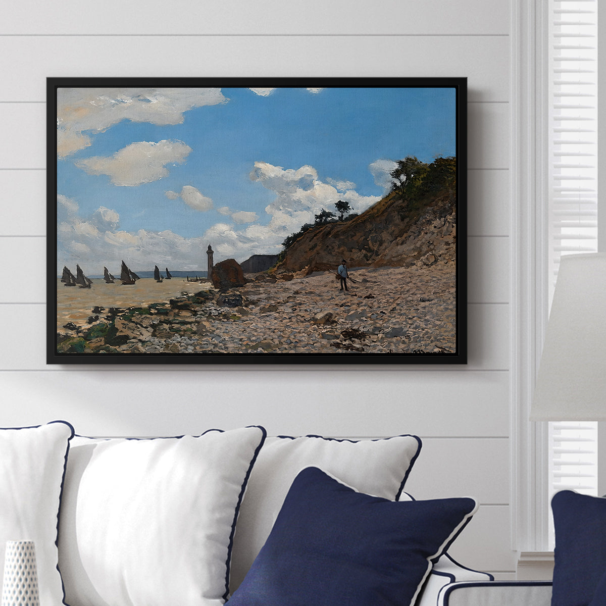 The Beach at Honfleur, 1864 by Claude Monet displayed above a sofa in a living room in a black floater frame, featuring an impressionist seaside with people and sailboats under bright sky.
