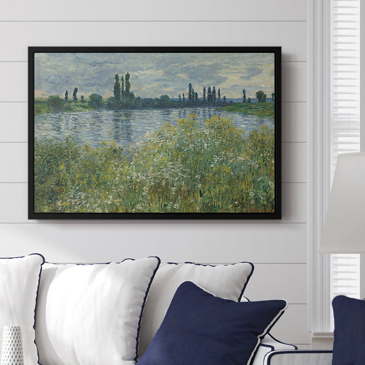 Banks of the Seine, Vétheuil, 1880 by Claude Monet displayed above a sofa in a living room in a black floater frame, featuring an impressionist view of Seine with reflections and greenery.