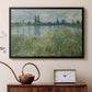 Black floater framed canvas featuring a tranquil riverside scene with houses and vegetation, displayed above a cabinet in a soft-toned living room.