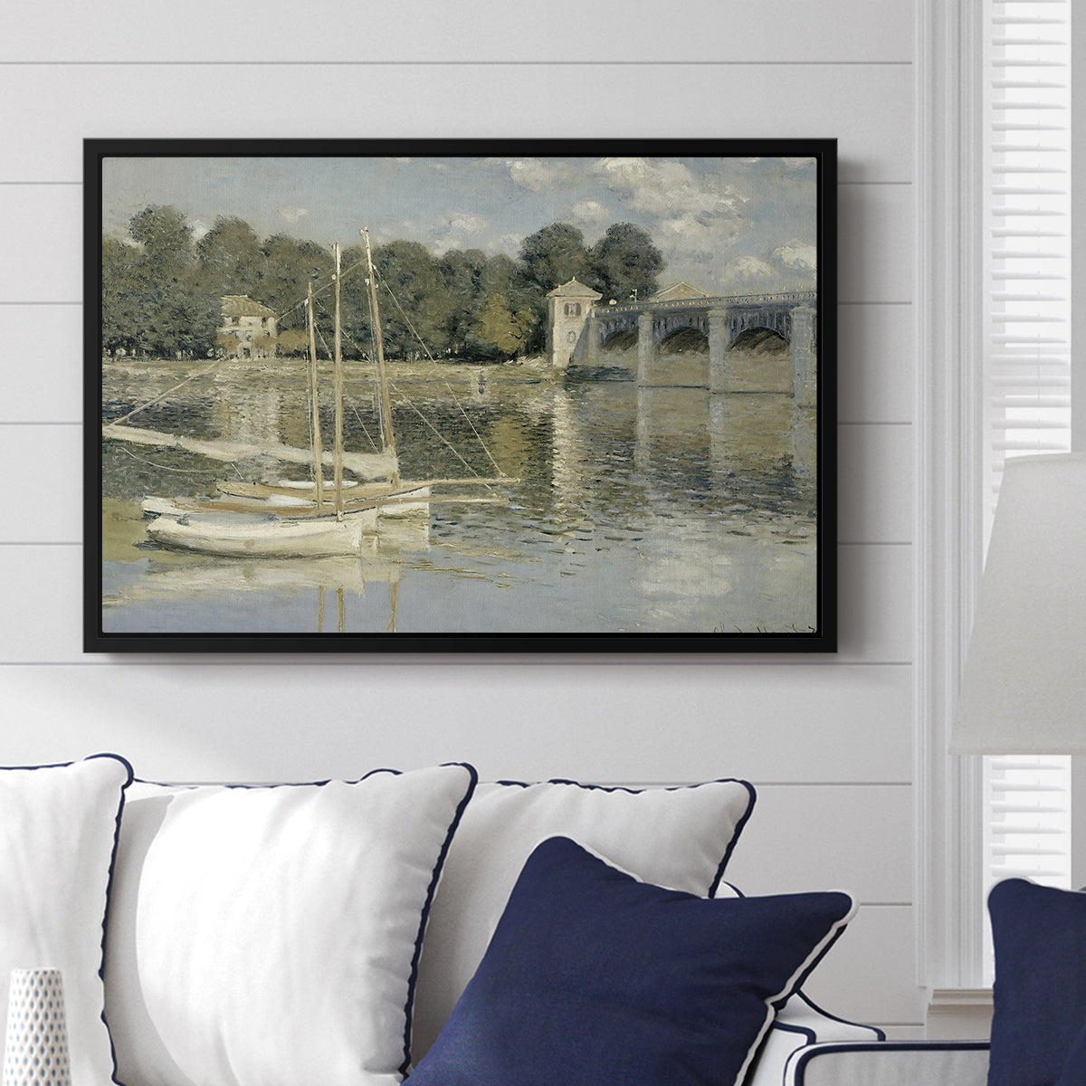 The Bridge at Argenteuil, 1874 by Claude Monet displayed above a sofa in a living room in a black floater frame, featuring an impressionist river scene with boats passing beneath bridge.
