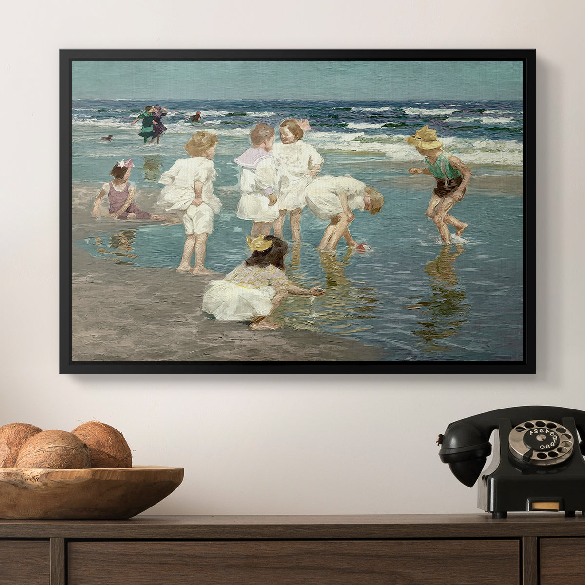 A Holiday, c. 1915 by Edward Henry Potthast in a black floater framed canvas displayed above a console table in a hallway, featuring a lively shoreline scene with people gathered in surf.