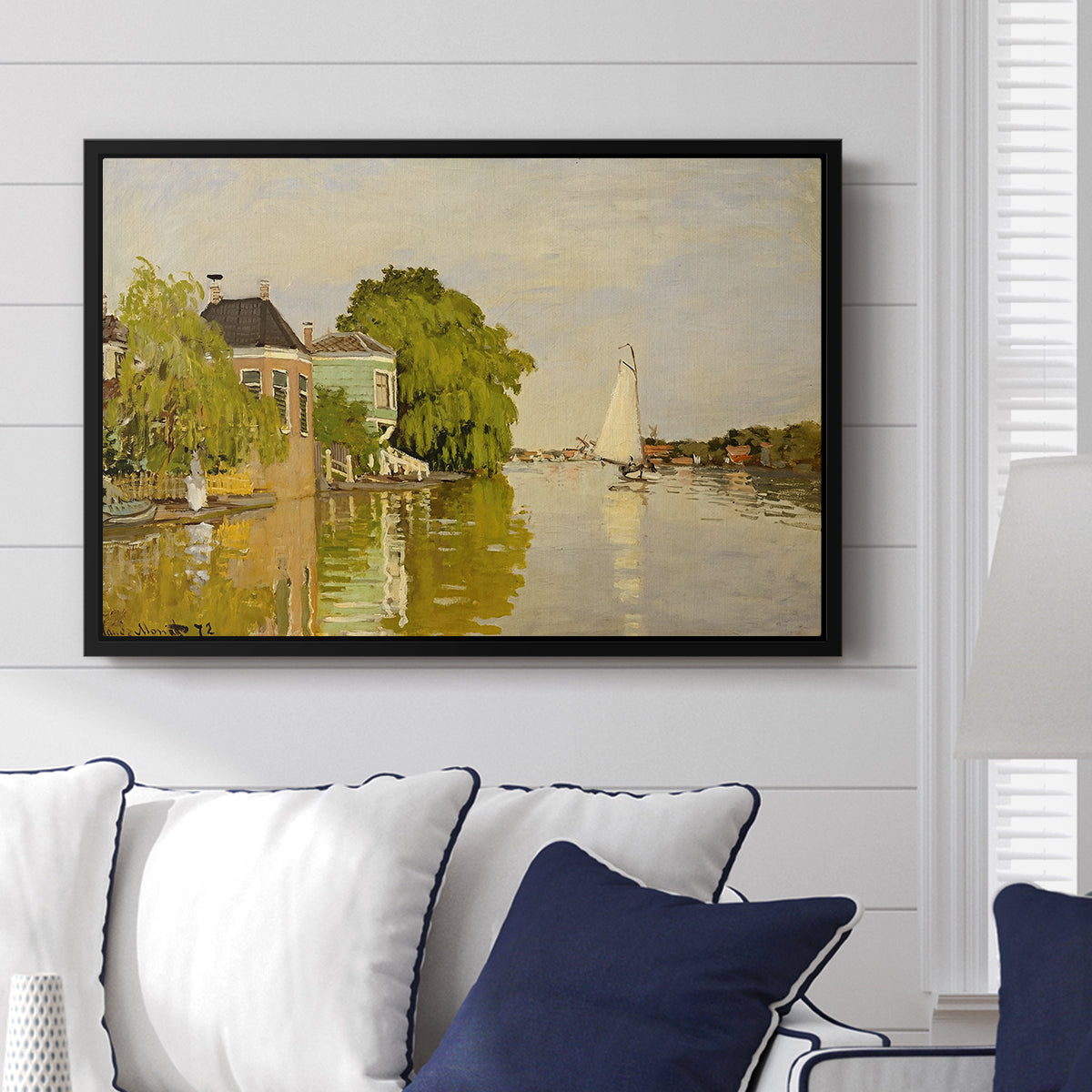 Houses on the Achterzaan, 1871 by Claude Monet displayed above a sofa in a living room in a black floater frame, featuring riverside homes with trees and mirrored reflections in water.
