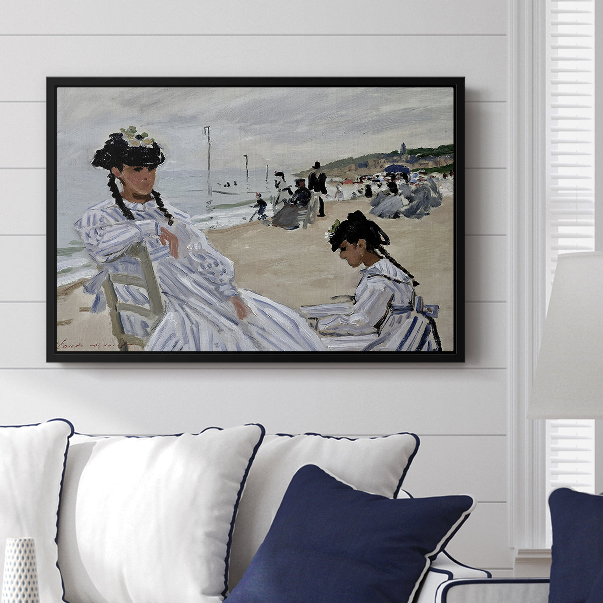 On the Beach at Trouville, 1870 by Claude Monet displayed above a sofa in a living room in a black floater frame, featuring an impressionist seaside scene with women in dresses and hats.