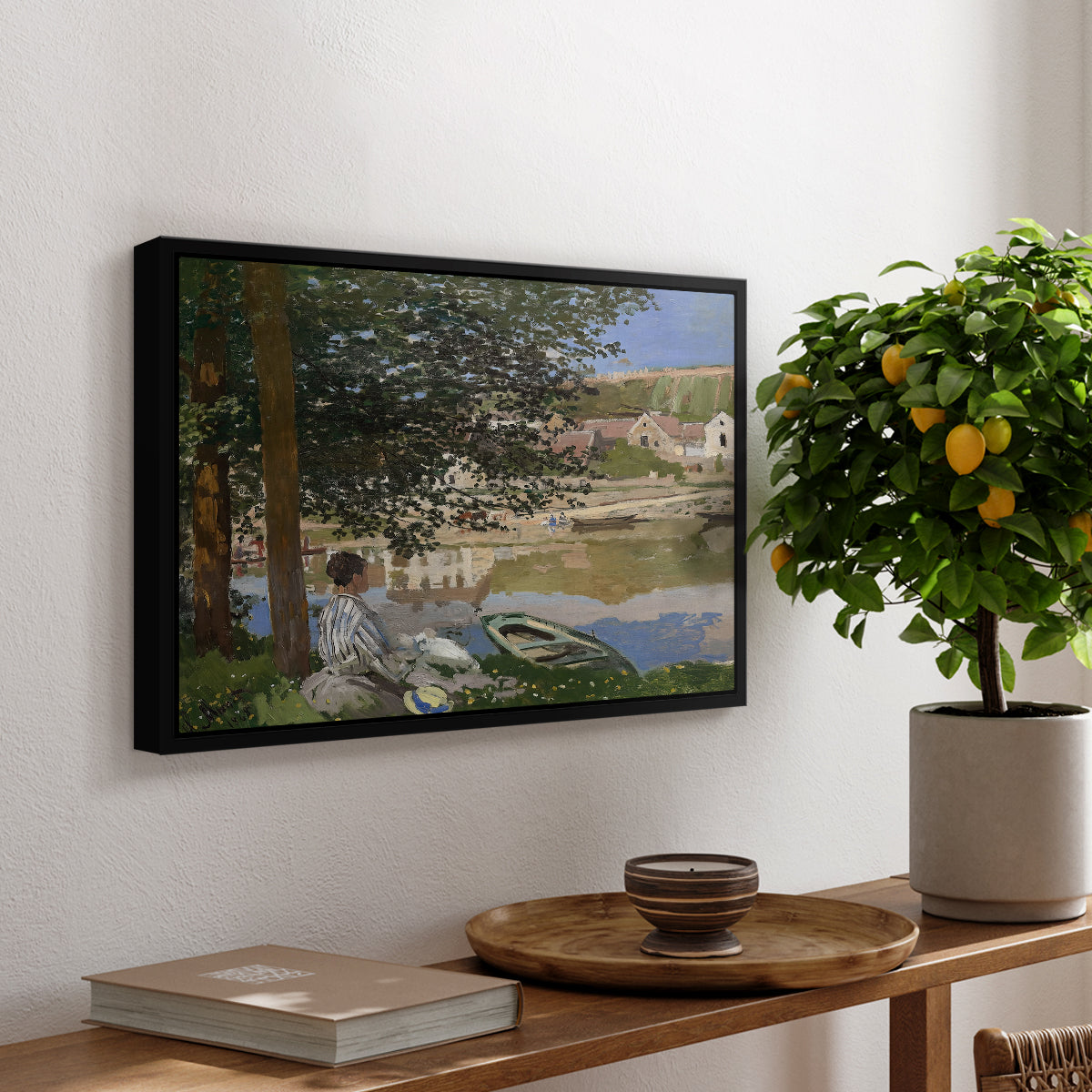 On the Bank of the Seine, Bennecourt, 1868 by Claude Monet, presented in a black floater framed canvas featuring a woman and child seated along riverbank beneath trees in soft daylight, displayed above a hallway console table.