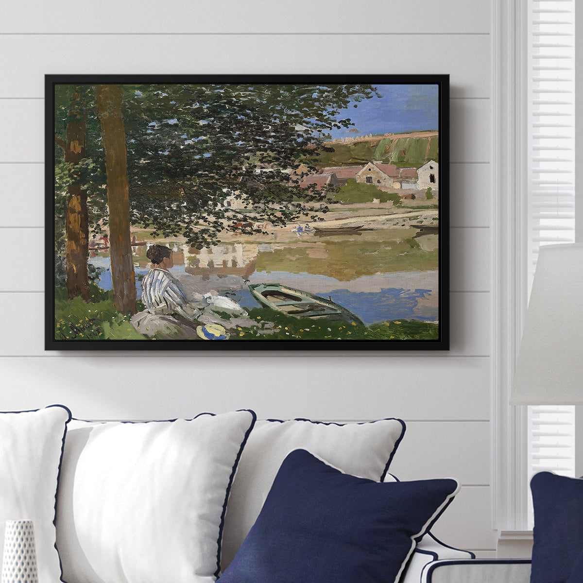 On the Bank of the Seine, Bennecourt, 1868 by Claude Monet displayed above a sofa in a living room in a black floater frame, featuring a peaceful riverside scene with figures resting in grassy area by water.