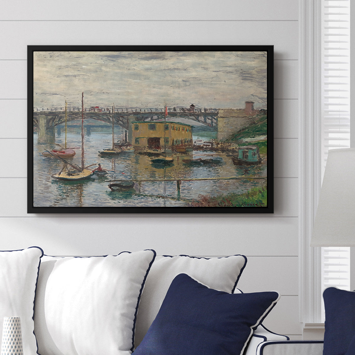 Bridge at Argenteuil on a Gray Day, c. 1876 by Claude Monet displayed above a sofa in a living room in a black floater frame, featuring an impressionist riverscape with overcast atmosphere and soft tones.