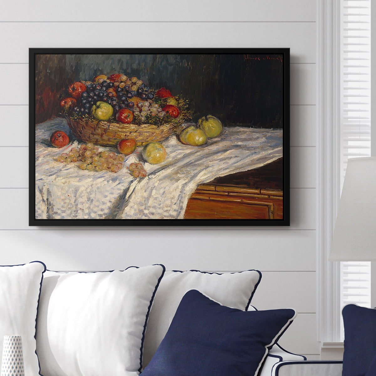Still Life with Apples and Grapes, 1880 by Claude Monet displayed above a sofa in a living room in a black floater frame, featuring an impressionist still life of fruit placed on cloth-covered surface.