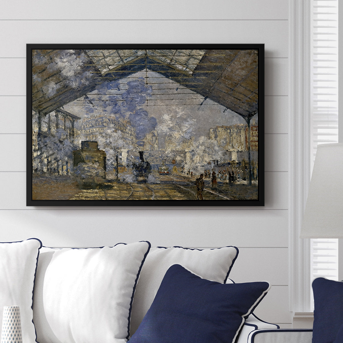The Gare Saint-Lazare, 1877 by Claude Monet displayed above a sofa in a living room in a black floater frame, featuring an impressionist scene of trains and smoke inside busy railway station.