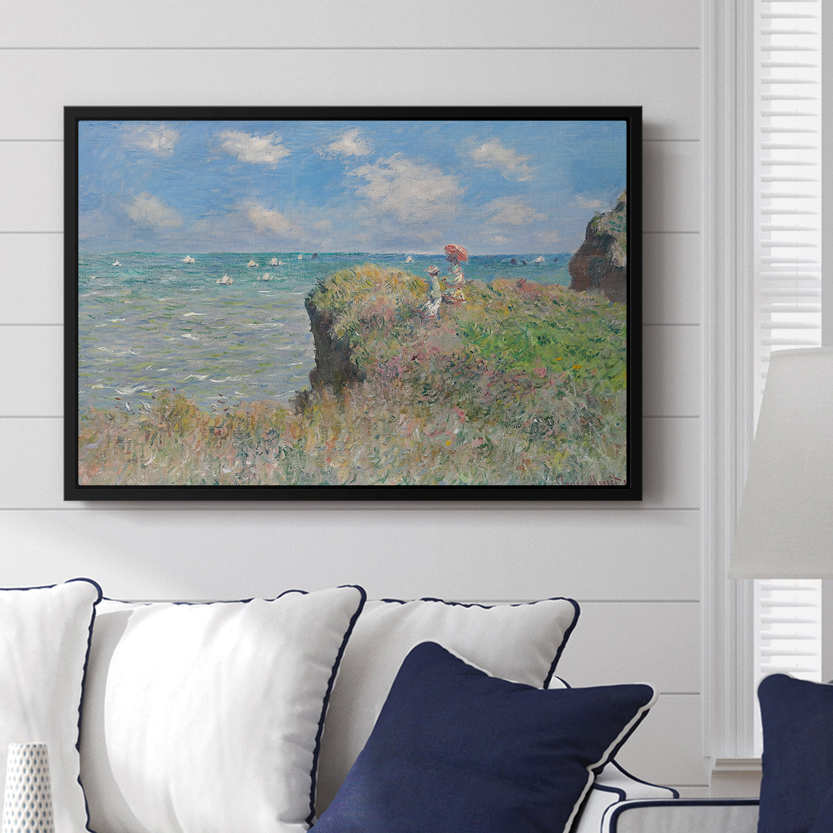 The Cliff Walk at Pourville, 1882 by Claude Monet displayed above a sofa in a living room in a black floater frame, featuring an impressionist seaside landscape with people strolling above water.