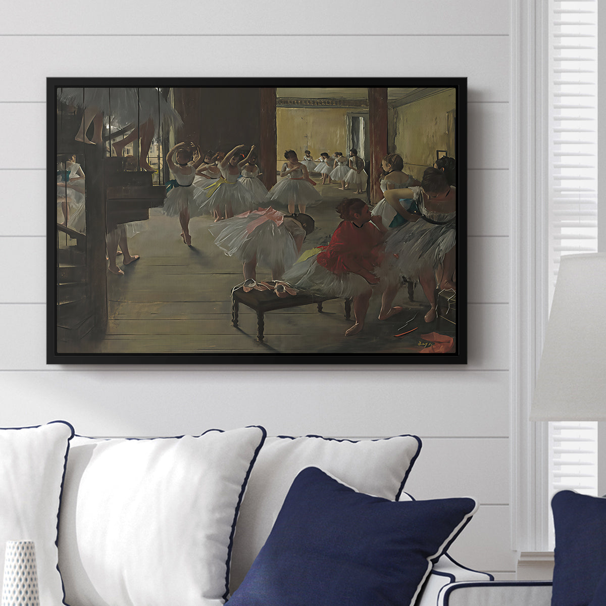 The Dance Class, c. 1873 by Edgar Degas displayed above a sofa in a living room in a black floater frame, featuring a rehearsal scene with dancers stretching and posing indoors.