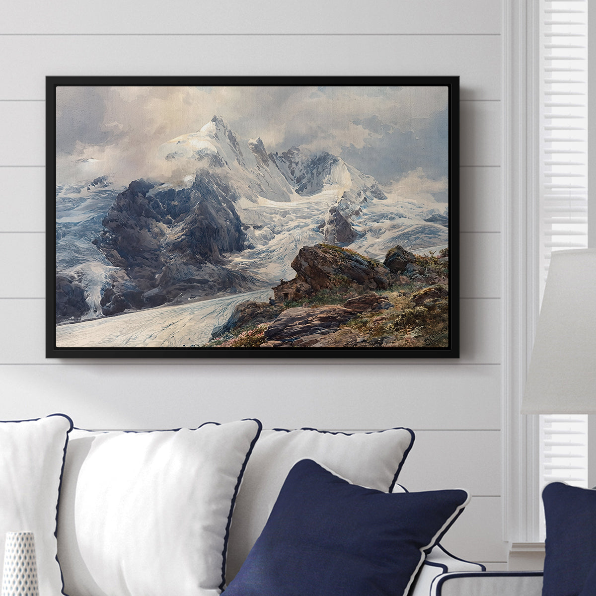 The Grossglockner, 1880 by Edward Theodore Compton displayed above a sofa in a living room in a black floater frame, featuring a dramatic mountain summit with snow and rugged slopes.