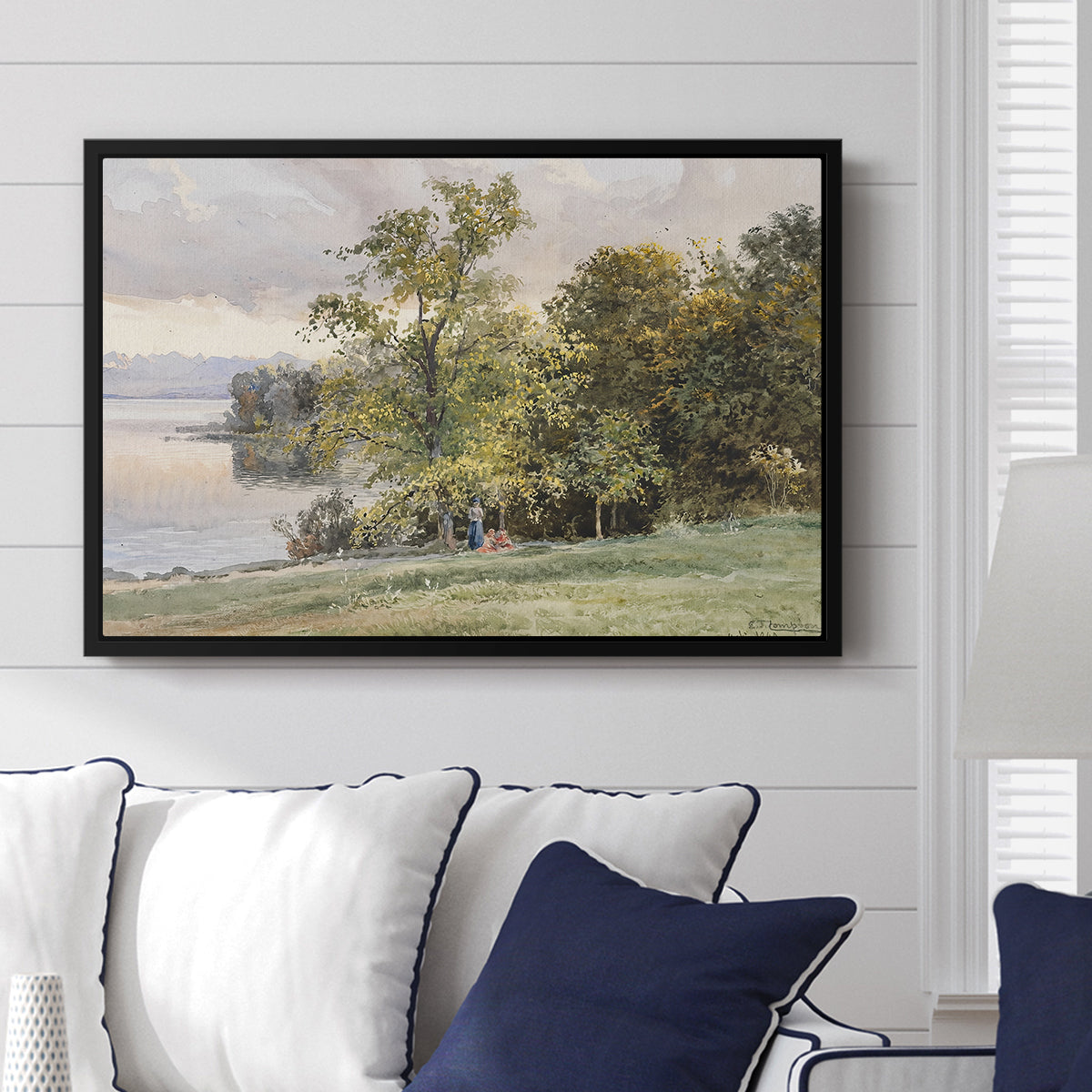 Landscape at Lake Starnberg, 1890 by Edward Theodore Compton displayed above a sofa in a living room in a black floater frame, featuring a peaceful lake scene with reflections and surrounding greenery.