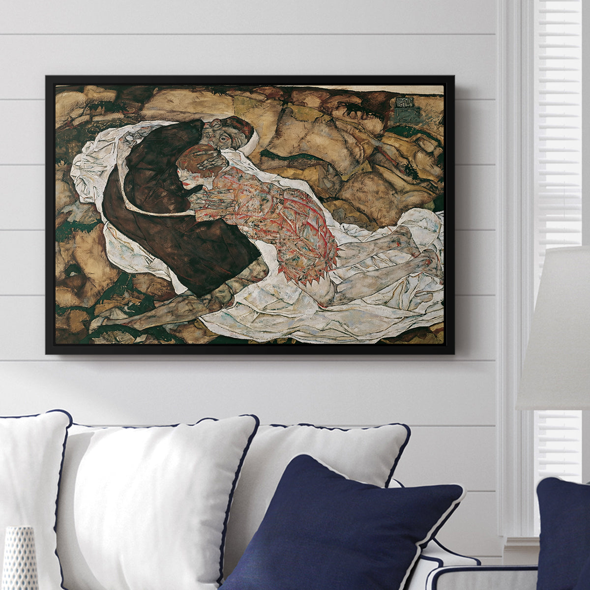 Death and the Maiden, 1915 by Egon Schiele displayed above a sofa in a living room in a black floater frame, featuring a symbolic scene of mortality with two figures clinging together.