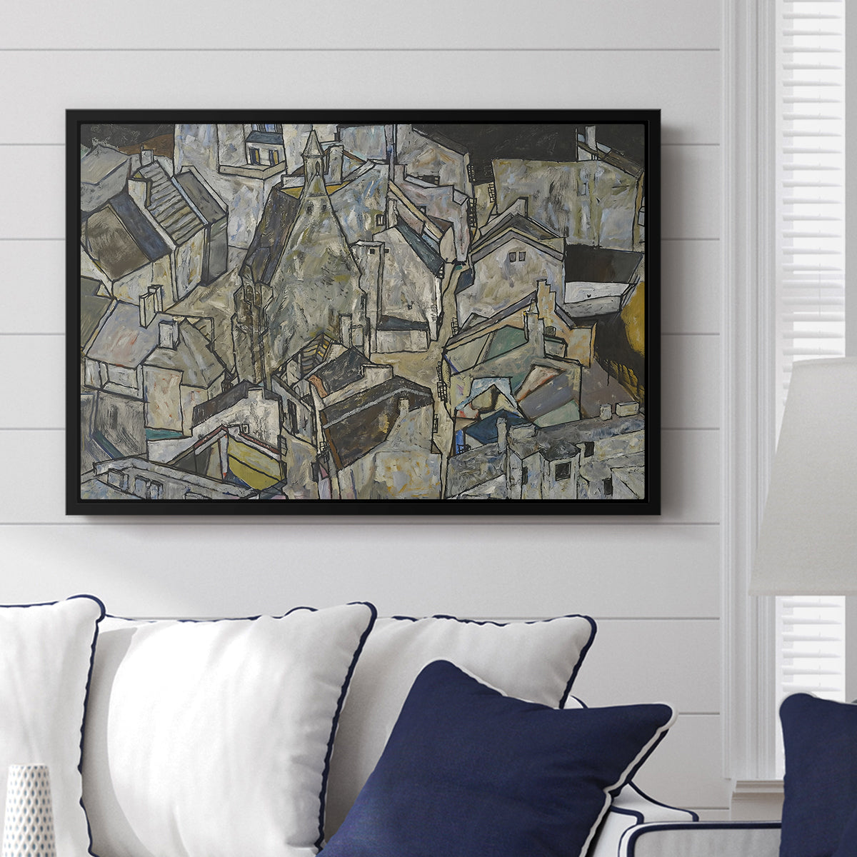 Mödling II, 1918 by Egon Schiele displayed above a sofa in a living room in a black floater frame, featuring an expressionist cityscape with tightly packed buildings and bold lines.