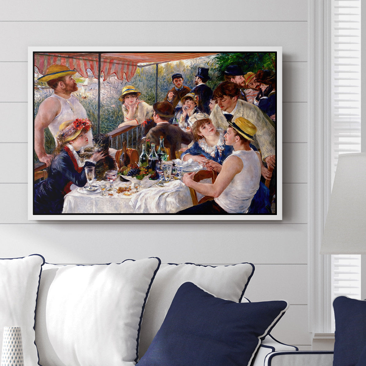Luncheon of the Boating Party (Le Déjeuner des canotiers), 1880-1881 by Pierre-Auguste Renoir displayed above a sofa in a living room in a white floater frame, featuring a lively gathering of men and women eating and socializing at outdoor table.