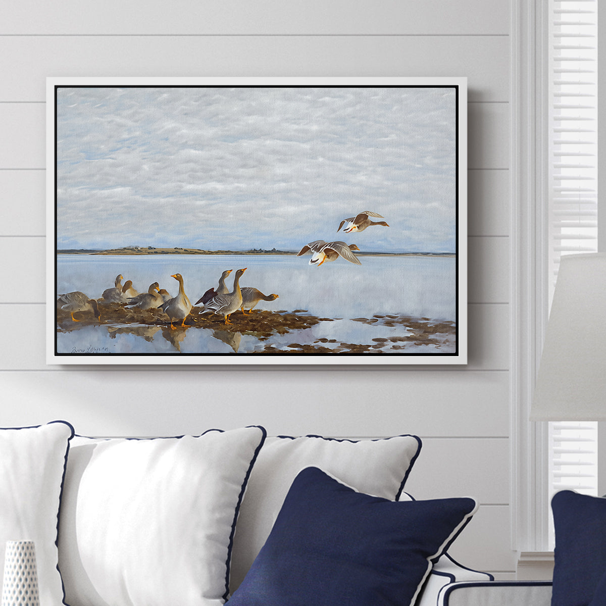 Greylag Geese (Grågäss), 1922 by Bruno Liljefors displayed above a sofa in a living room in a white floater frame, featuring birds standing and resting along shoreline with soft grass and water.