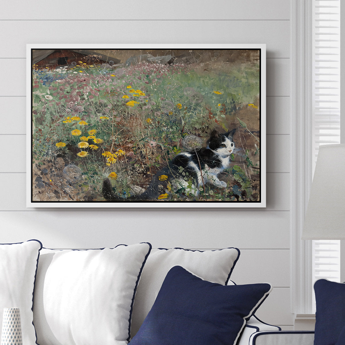 Cat on a Flowery Meadow, 1887 by Bruno Liljefors displayed above a sofa in a living room in a white floater frame, featuring a small cat resting among colorful flowers in grassy field.