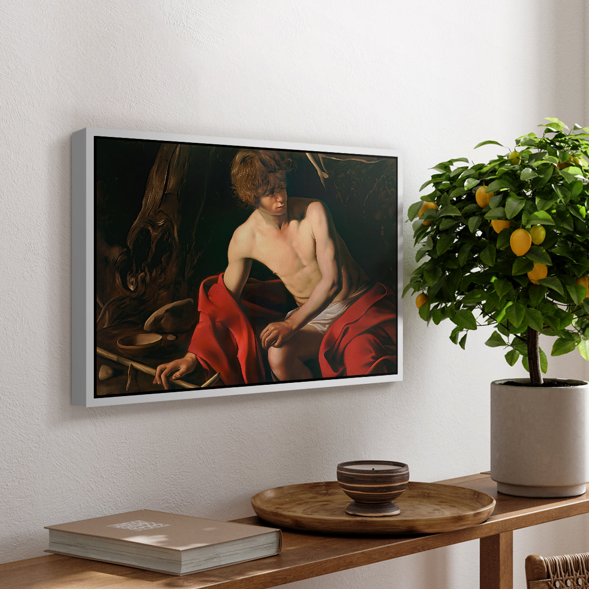 Saint John the Baptist in the Wilderness, c. 1603-1604 by Caravaggio, presented in a white floater framed canvas featuring a young Saint John seated in wilderness with staff and red drapery, displayed above a hallway console table.