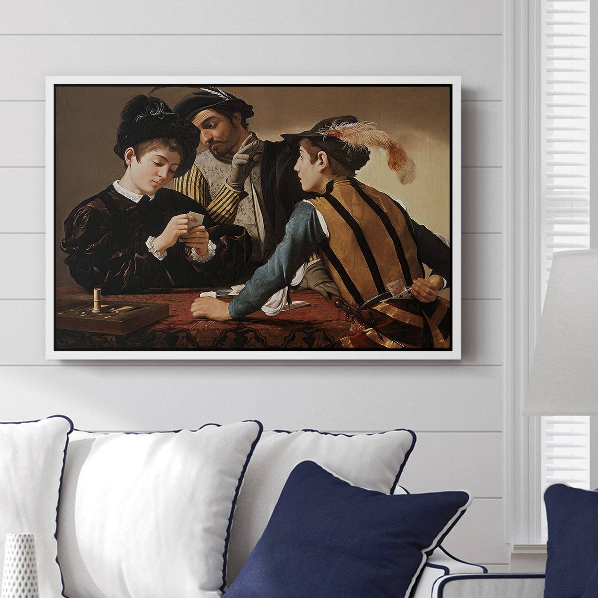 The Cardsharps, c. 1594 by Caravaggio displayed above a sofa in a living room in a white floater frame, featuring a genre scene of card game with deception and hidden accomplice.