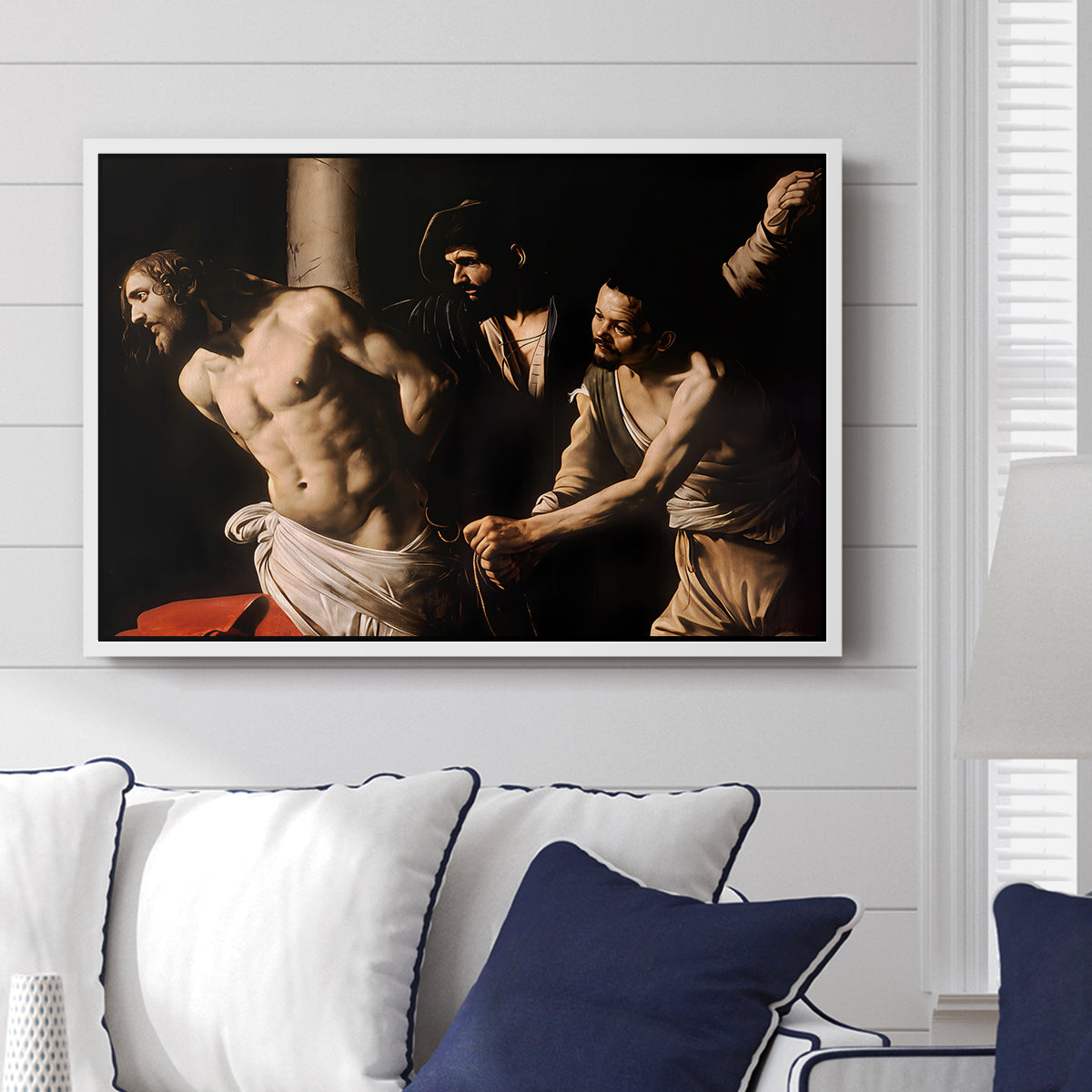 The Flagellation of Christ at the Column (Rouen Version), 1606 by Caravaggio displayed above a sofa in a living room in a white floater frame, featuring a dramatic religious scene of Christ enduring flagellation in dark interior.