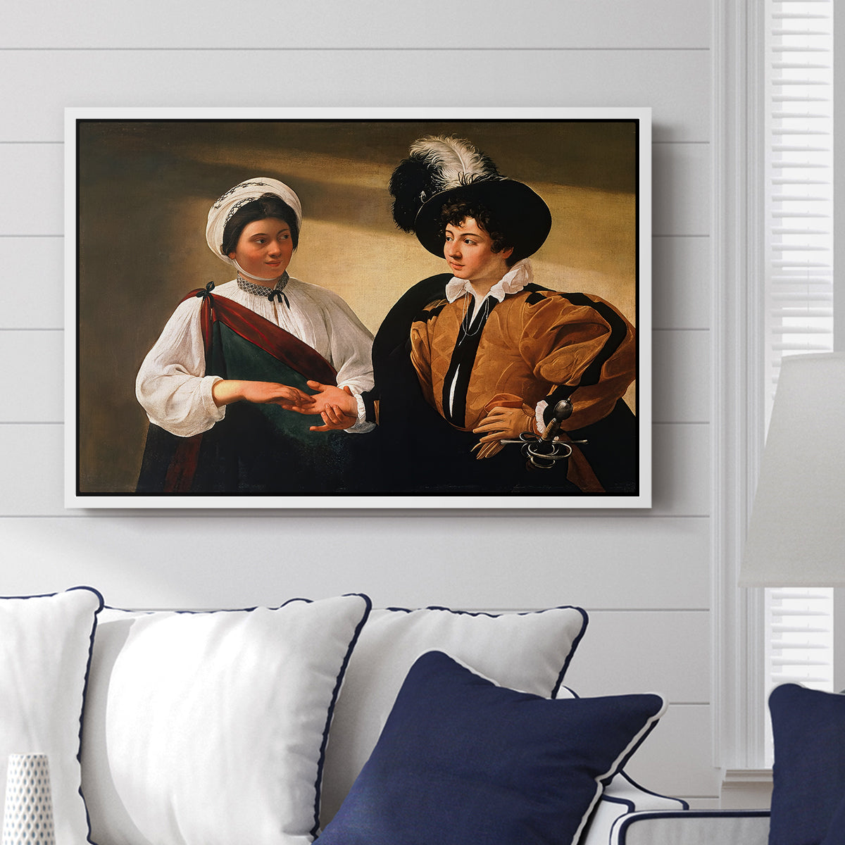 The Fortune Teller (Louvre Version), c. 1595 by Caravaggio displayed above a sofa in a living room in a white floater frame, featuring a fortune teller examining hand of man as she slyly removes his ring.