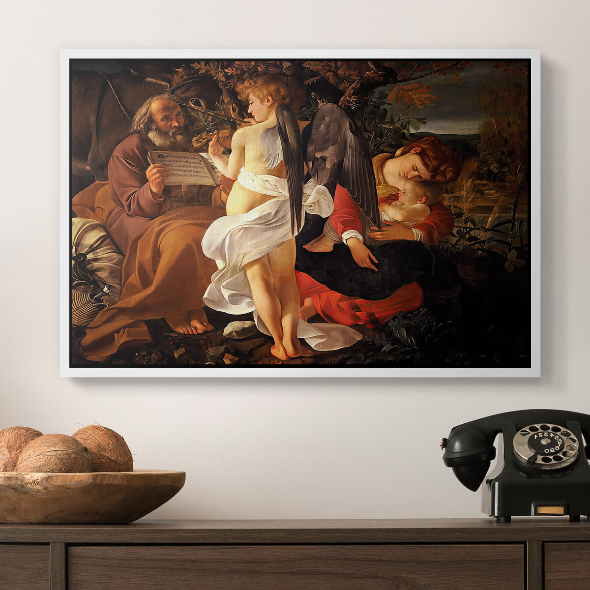 Rest on the Flight into Egypt, c. 1597 by Caravaggio in a white floater framed canvas displayed above a console table in a hallway, featuring a Baroque landscape with angel playing music for resting travelers.