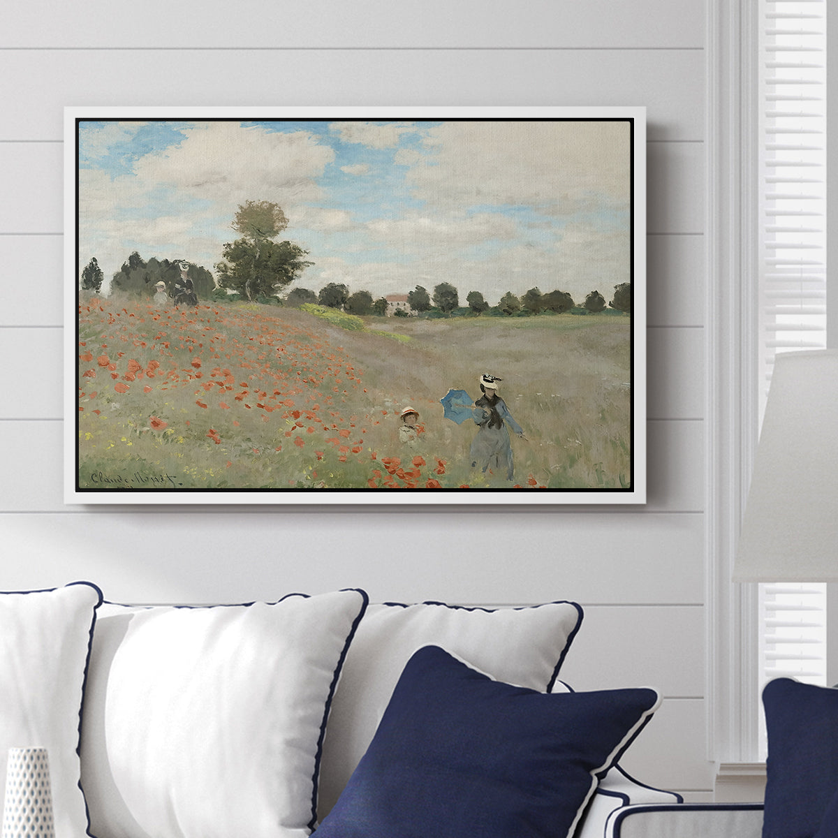 The Poppy Field near Argenteuil, 1873 by Claude Monet displayed above a sofa in a living room in a white floater frame, featuring an impressionist countryside scene with flowers and strolling figures.
