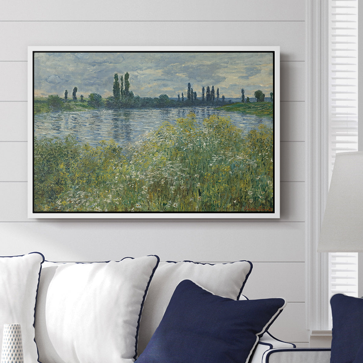 Banks of the Seine, Vétheuil, 1880 by Claude Monet displayed above a sofa in a living room in a white floater frame, featuring an impressionist view of Seine with reflections and greenery.