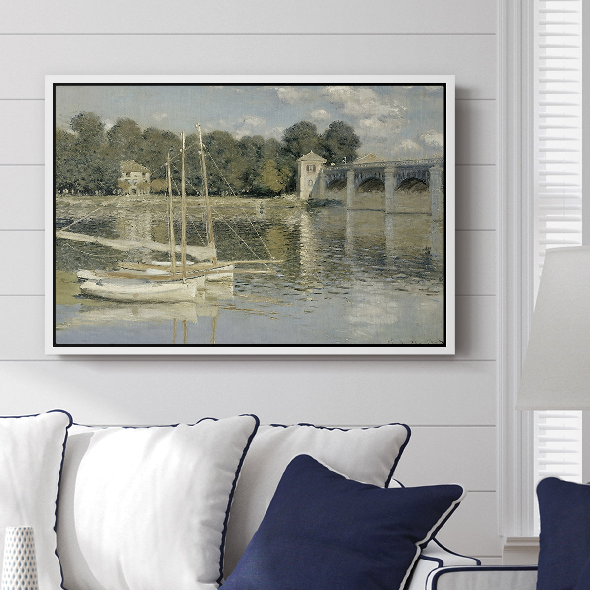 The Bridge at Argenteuil, 1874 by Claude Monet displayed above a sofa in a living room in a white floater frame, featuring an impressionist river scene with boats passing beneath bridge.