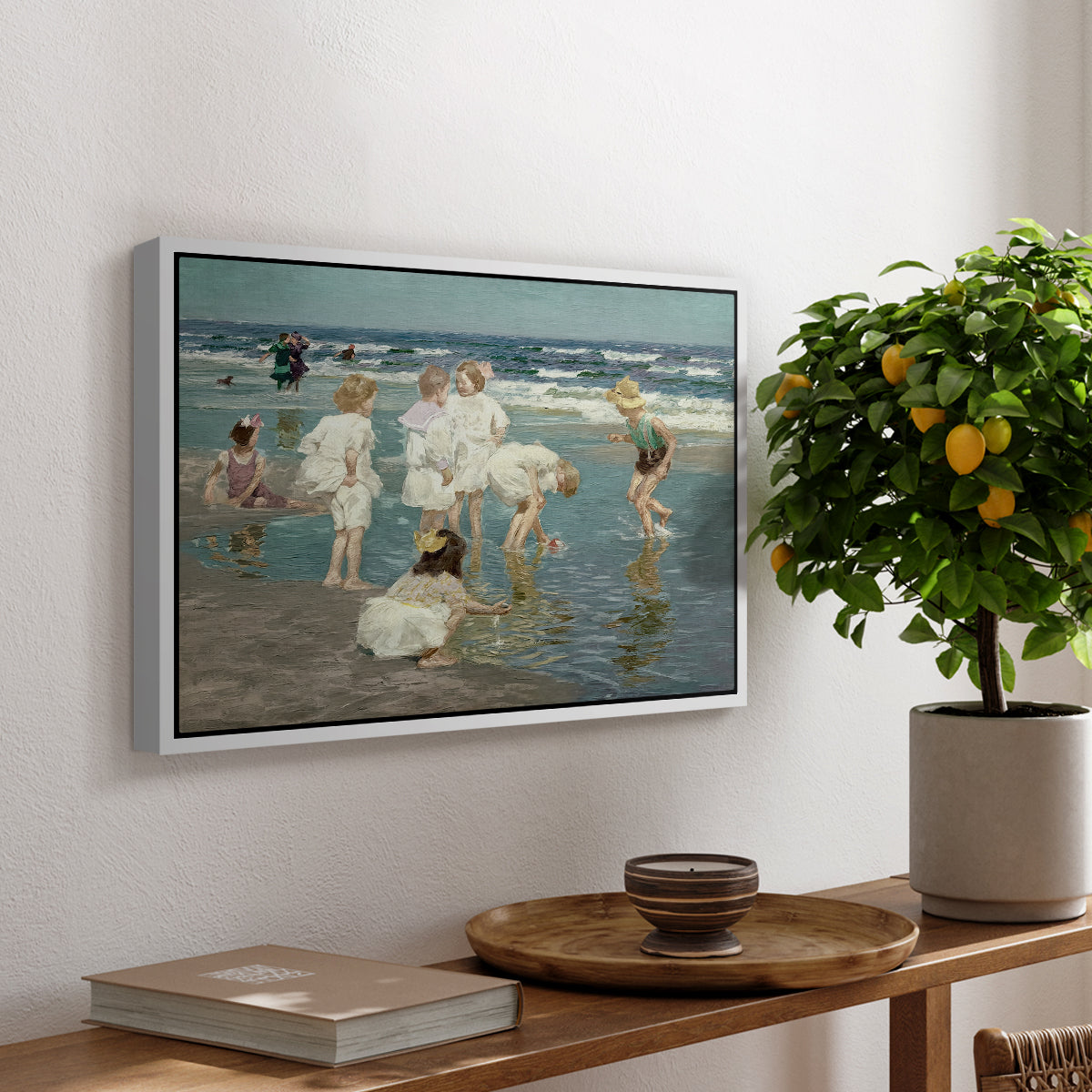 A Holiday, c. 1915 by Edward Henry Potthast, presented in a white floater framed canvas featuring people relaxing and wading in shallow ocean water along a sunny beach, displayed above a hallway console table.
