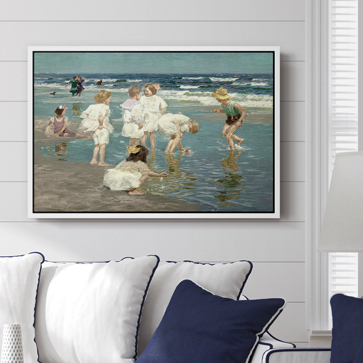 A Holiday, c. 1915 by Edward Henry Potthast displayed above a sofa in a living room in a white floater frame, featuring a beach scene with figures bathing and standing in waves under bright sky.