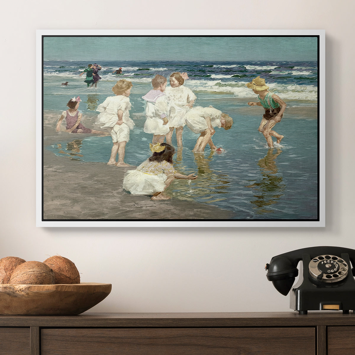 A Holiday, c. 1915 by Edward Henry Potthast in a white floater framed canvas displayed above a console table in a hallway, featuring a lively shoreline scene with people gathered in surf.