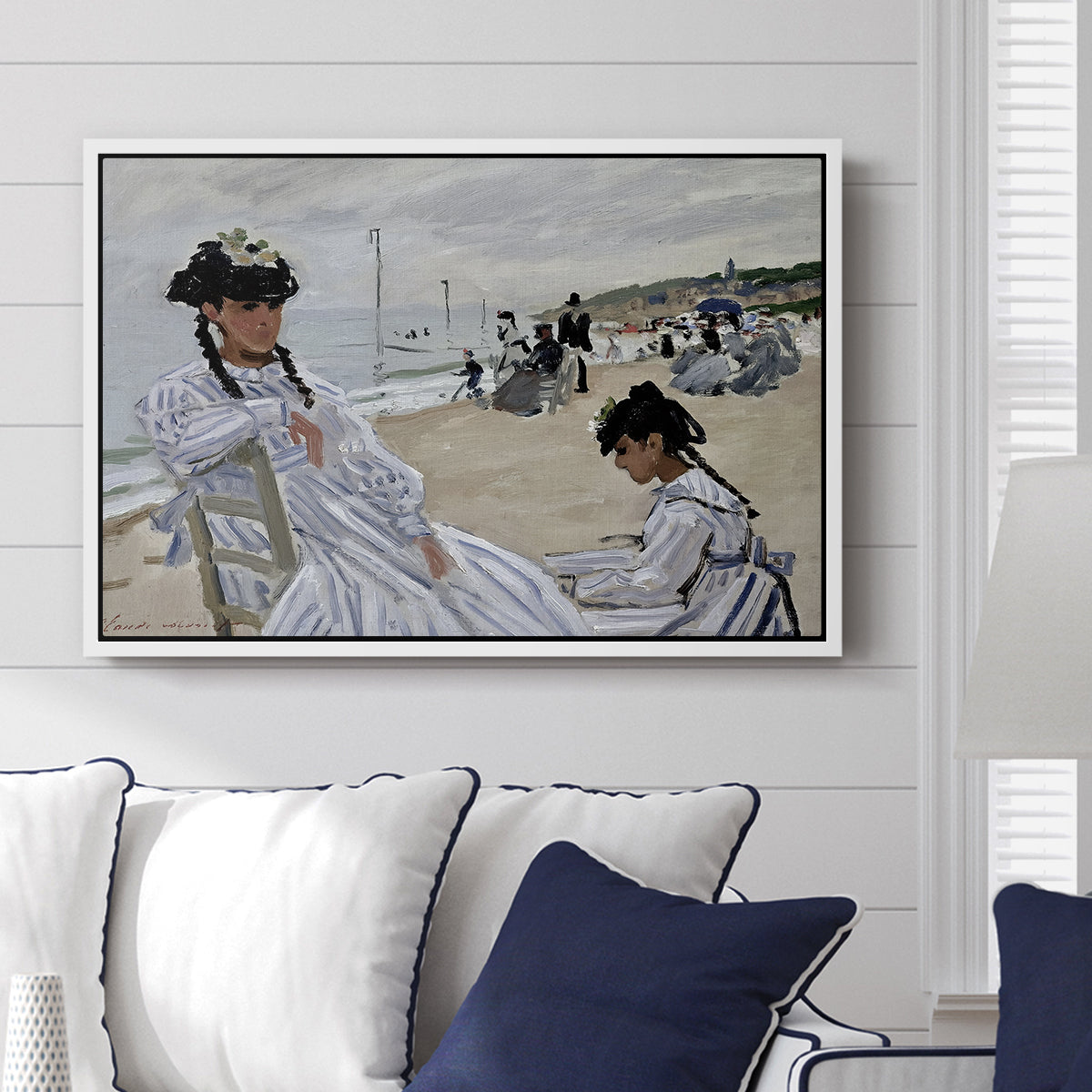 On the Beach at Trouville, 1870 by Claude Monet displayed above a sofa in a living room in a white floater frame, featuring an impressionist seaside scene with women in dresses and hats.