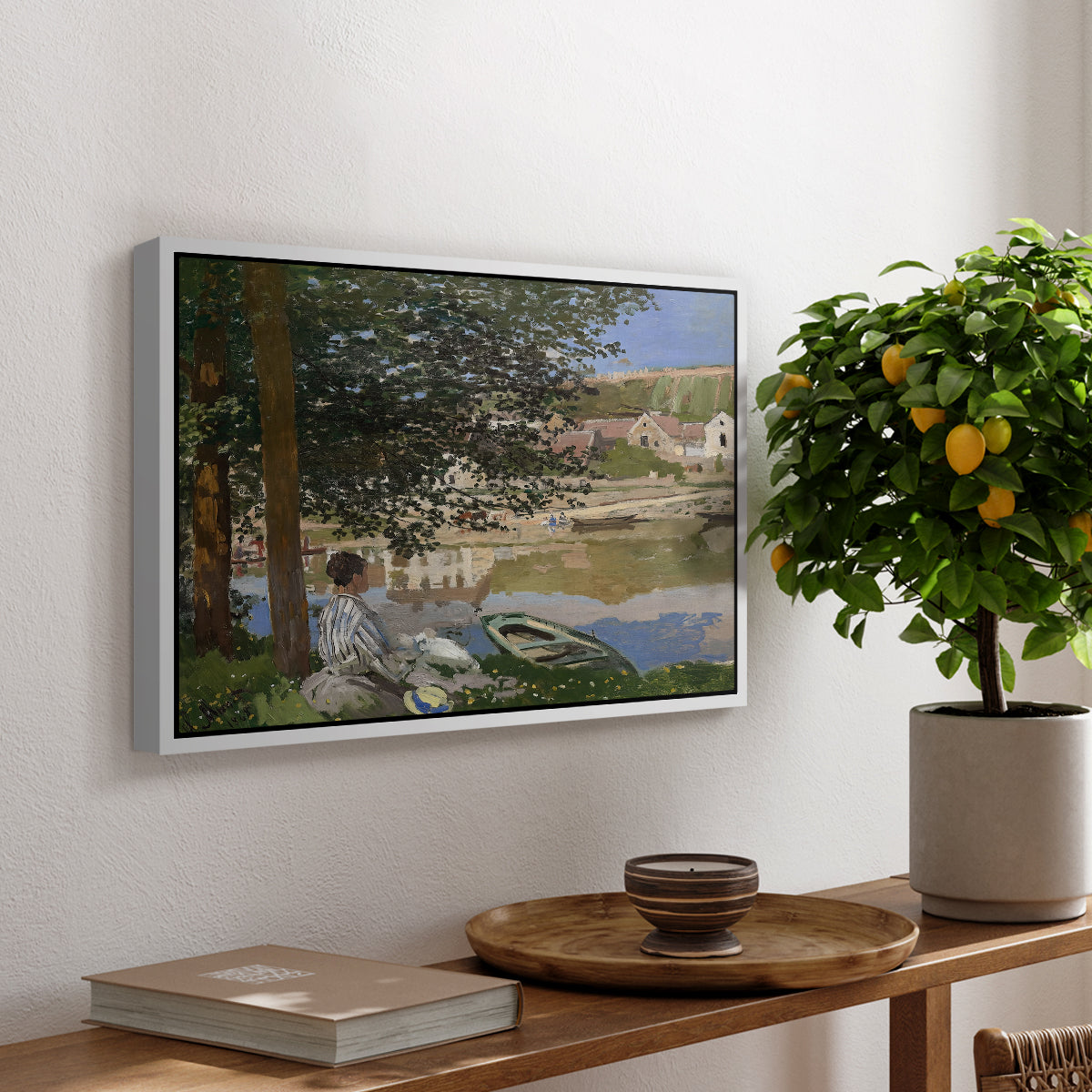 On the Bank of the Seine, Bennecourt, 1868 by Claude Monet, presented in a white floater framed canvas featuring a woman and child seated along riverbank beneath trees in soft daylight, displayed above a hallway console table.