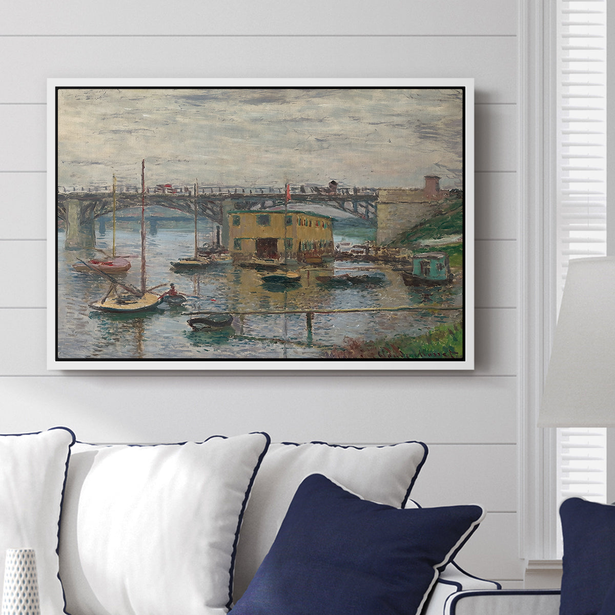 Bridge at Argenteuil on a Gray Day, c. 1876 by Claude Monet displayed above a sofa in a living room in a white floater frame, featuring an impressionist riverscape with overcast atmosphere and soft tones.