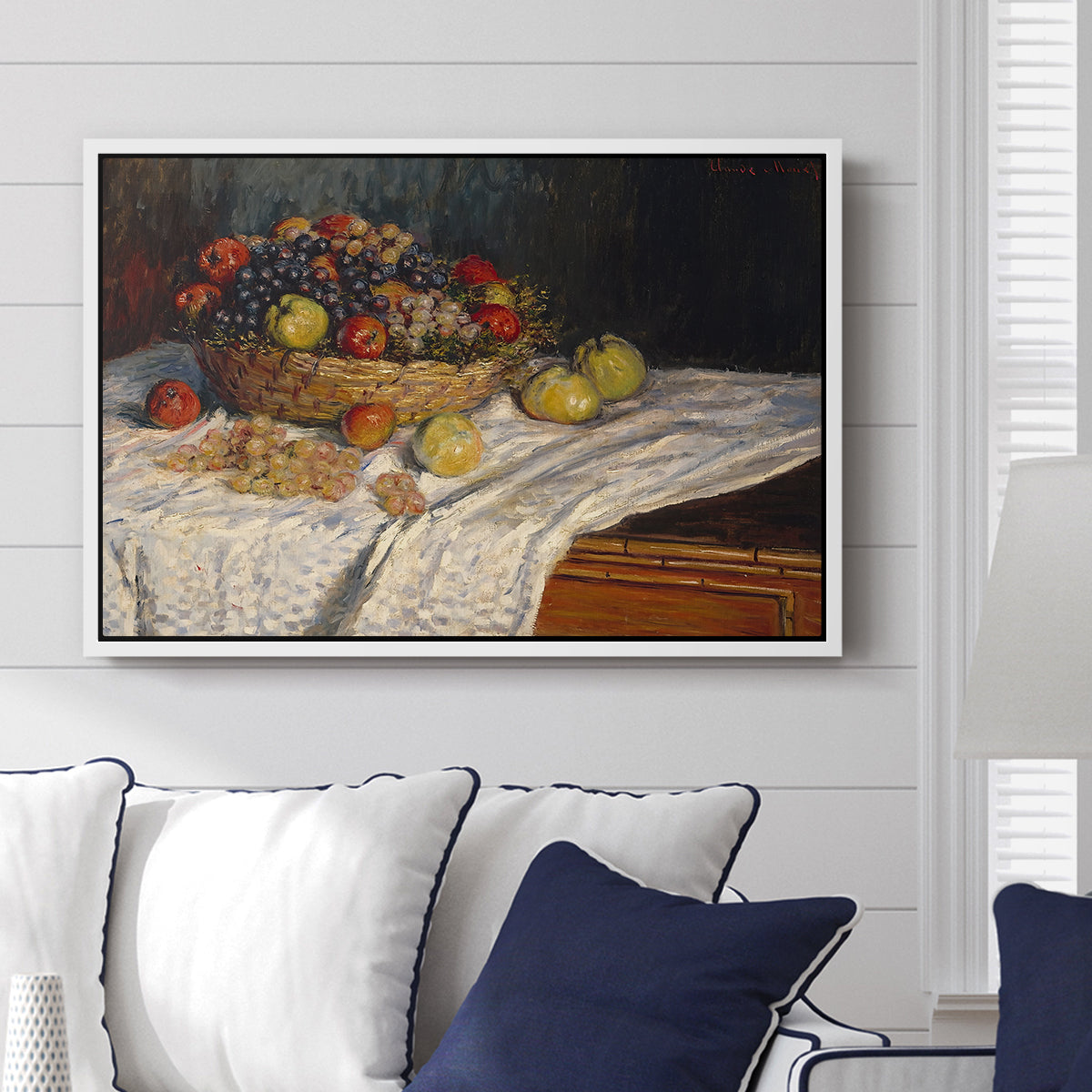 Still Life with Apples and Grapes, 1880 by Claude Monet displayed above a sofa in a living room in a white floater frame, featuring an impressionist still life of fruit placed on cloth-covered surface.