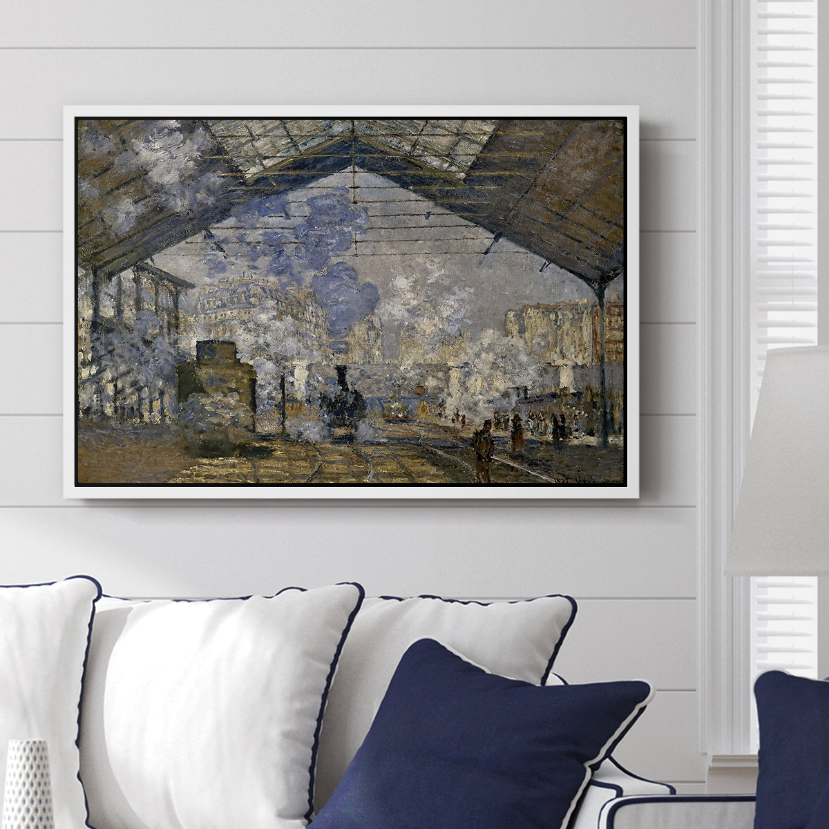 The Gare Saint-Lazare, 1877 by Claude Monet displayed above a sofa in a living room in a white floater frame, featuring an impressionist scene of trains and smoke inside busy railway station.