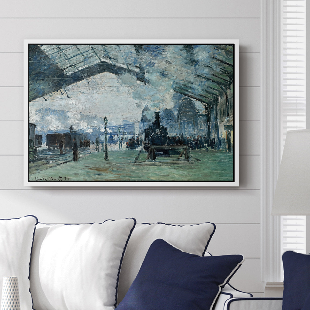 The Saint-Lazare Station, 1877 by Claude Monet displayed above a sofa in a living room in a white floater frame, featuring an impressionist railway scene with locomotives and haze.