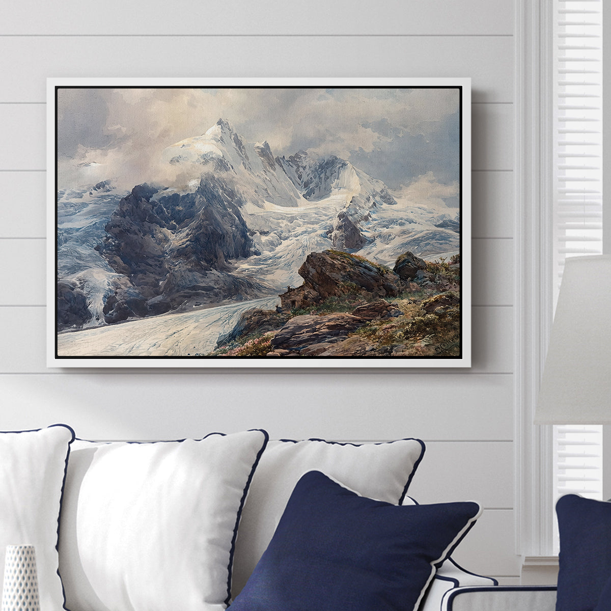The Grossglockner, 1880 by Edward Theodore Compton displayed above a sofa in a living room in a white floater frame, featuring a dramatic mountain summit with snow and rugged slopes.