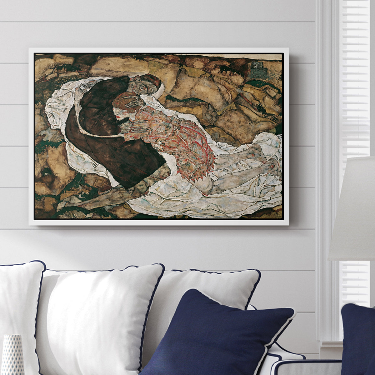 Death and the Maiden, 1915 by Egon Schiele displayed above a sofa in a living room in a white floater frame, featuring a symbolic scene of mortality with two figures clinging together.