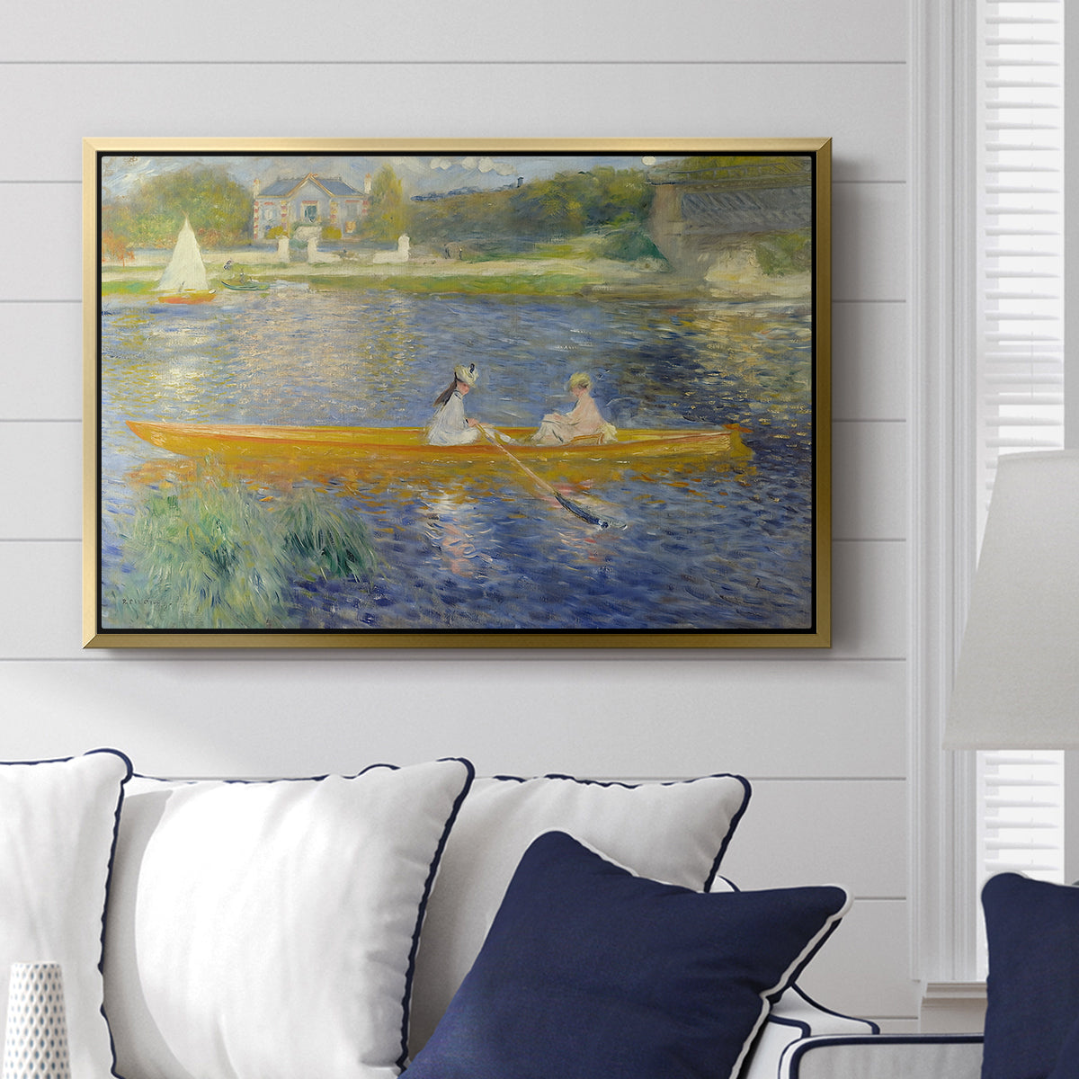 The Skiff (La Yole), 1875 by Pierre-Auguste Renoir displayed above a sofa in a living room in a gold floater frame, featuring an impressionist boating scene with people relaxing in a narrow skiff.