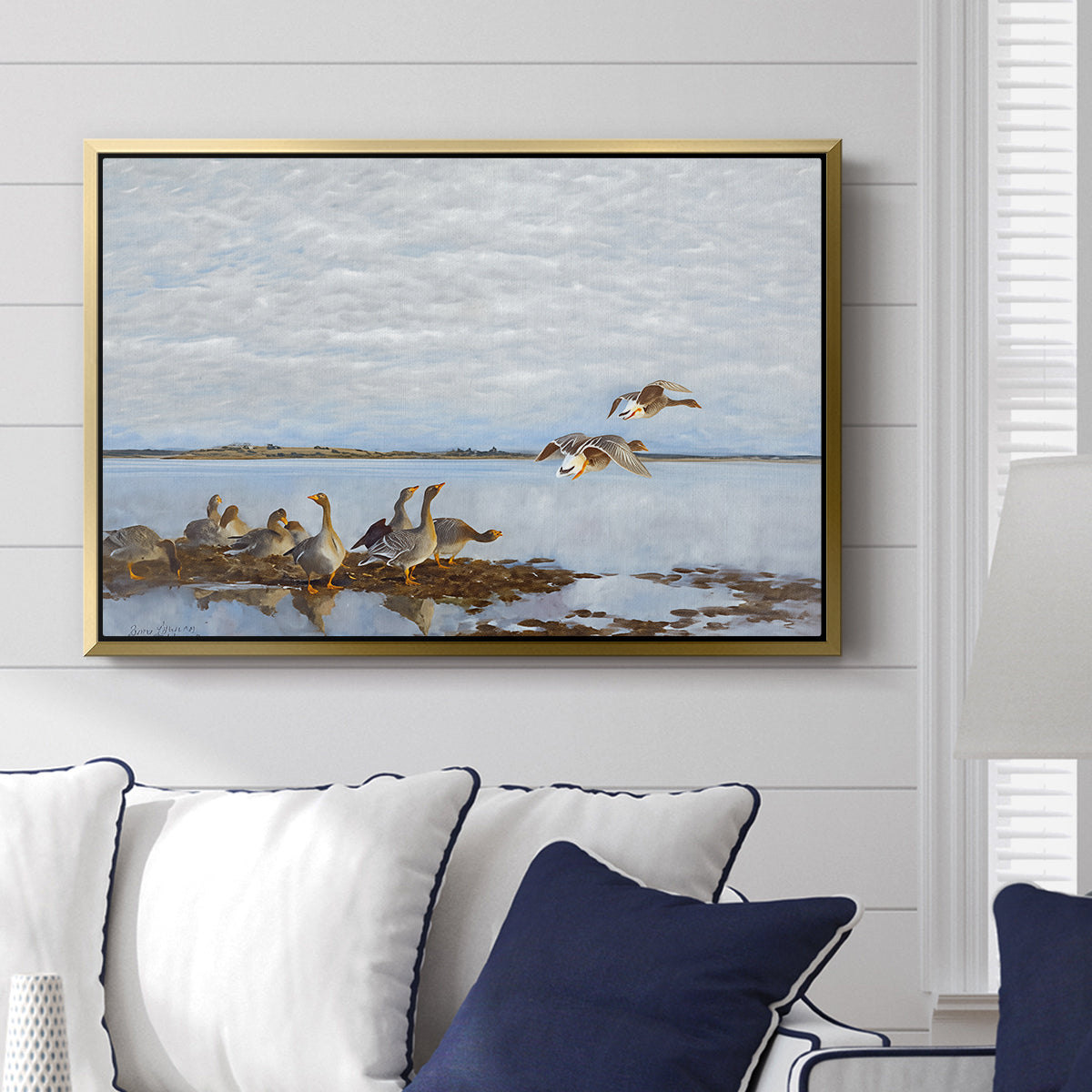 Greylag Geese (Grågäss), 1922 by Bruno Liljefors displayed above a sofa in a living room in a gold floater frame, featuring birds standing and resting along shoreline with soft grass and water.