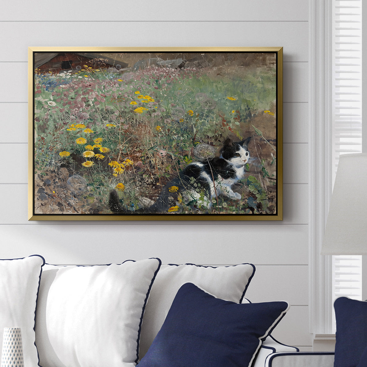 Cat on a Flowery Meadow, 1887 by Bruno Liljefors displayed above a sofa in a living room in a gold floater frame, featuring a small cat resting among colorful flowers in grassy field.