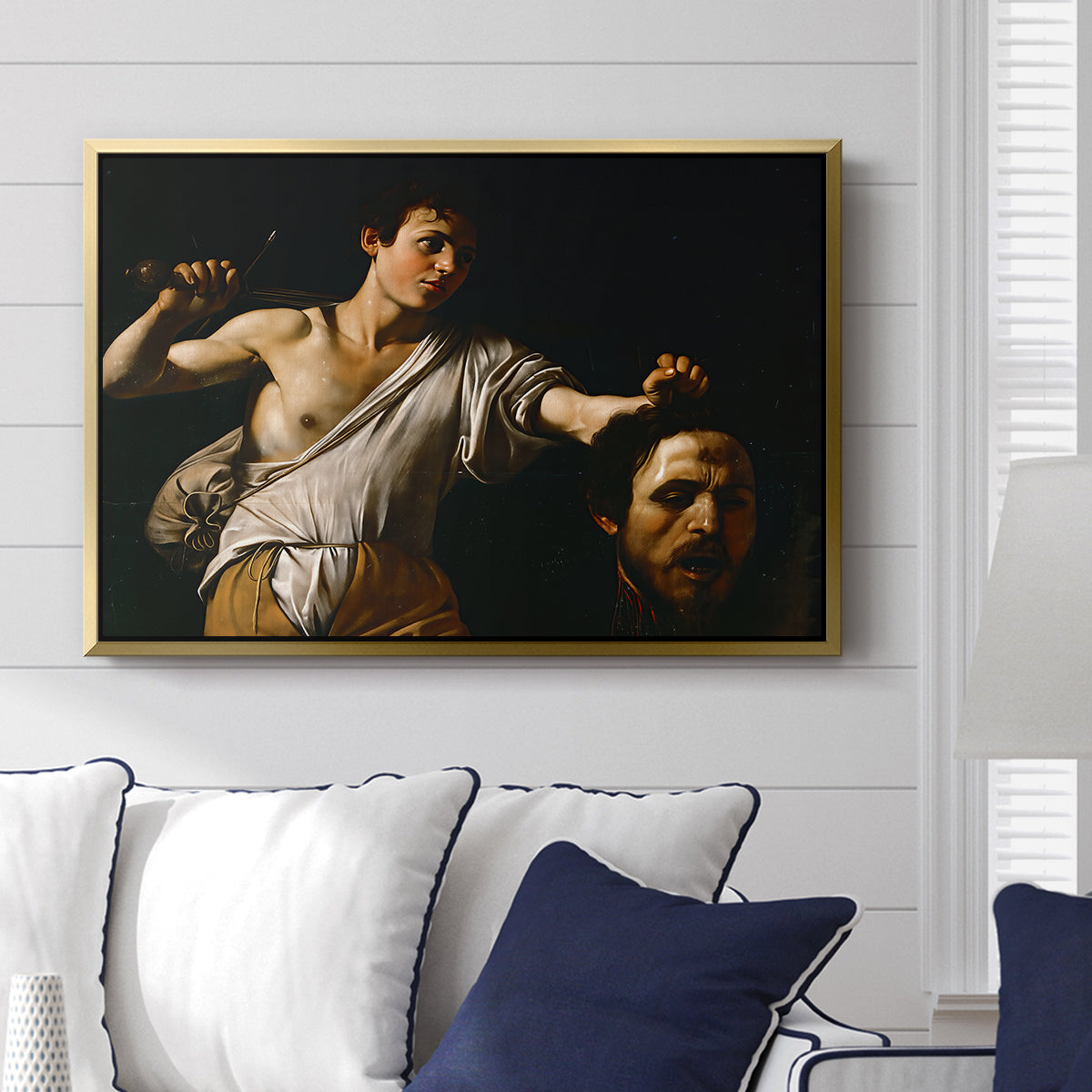 David with the Head of Goliath (Vienna Version), c. 1607 by Caravaggio displayed above a sofa in a living room in a gold floater frame, featuring a biblical scene of David gripping Goliath's head with dramatic lighting.
