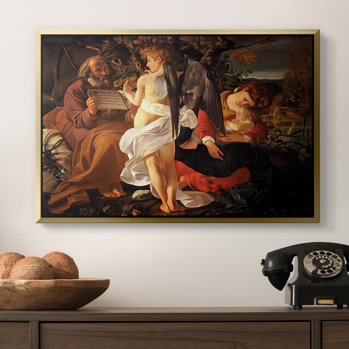 Rest on the Flight into Egypt, c. 1597 by Caravaggio in a gold floater framed canvas displayed above a console table in a hallway, featuring a Baroque landscape with angel playing music for resting travelers.