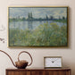Gold floater framed canvas featuring a tranquil riverside scene with houses and vegetation, displayed above a cabinet in a soft-toned living room.
